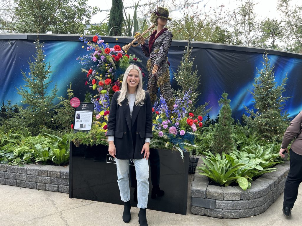 Emily Ilchena, the owner and floral designer at Juniper Jayne Creative stands in front of her piece at The Leaf on Jan. 16, 2025. The floral sculpture, which is made up of hundreds of flowers is of a woman playing a trumpet. 